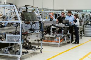 A manufacturing facility interior with a small leadership team standing near production equipment, reviewing revenue and performance charts, representing a company evaluating whether steady output equals real stability.