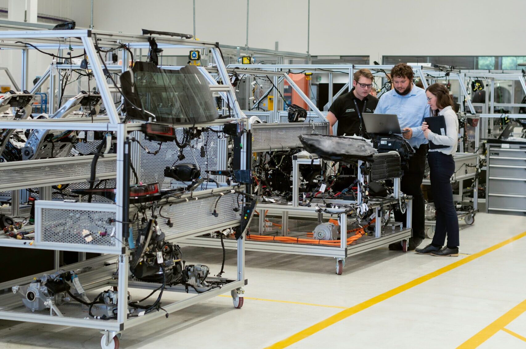 Manufacturing plant floor with a leadership team reviewing performance trends A manufacturing facility interior with a small leadership team standing near production equipment, reviewing revenue and performance charts, representing a company evaluating whether steady output equals real stability.